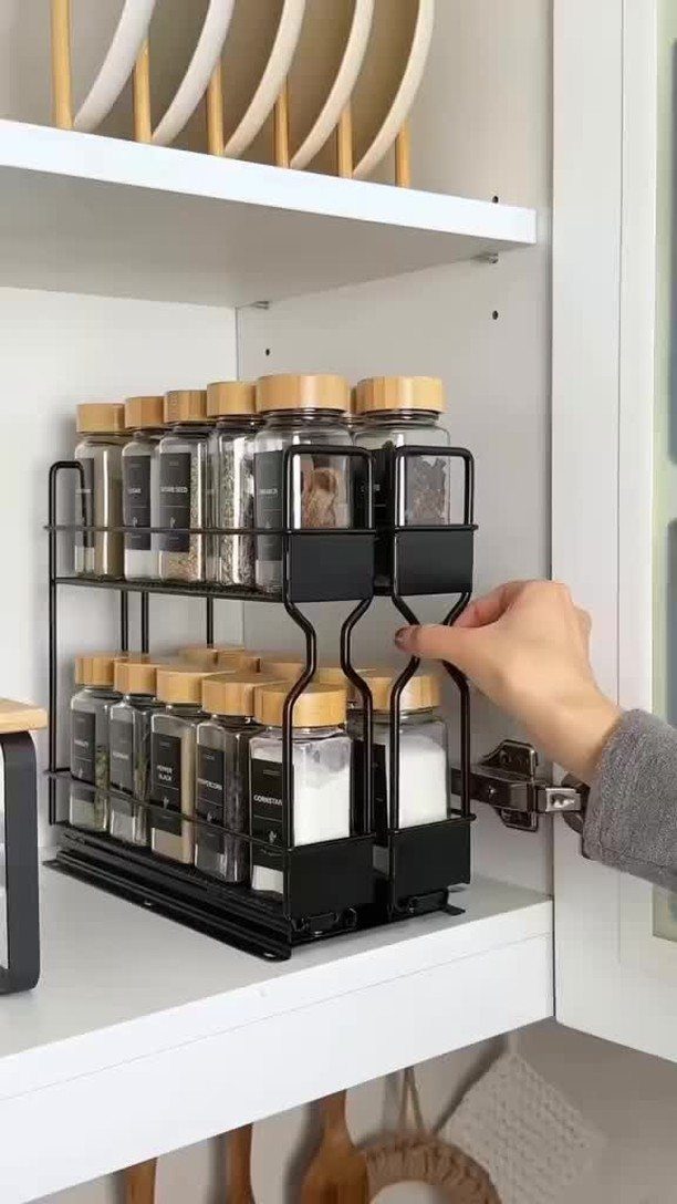 A hand reaches for a spice jar on a black two-tier metal rack inside a white cabinet.