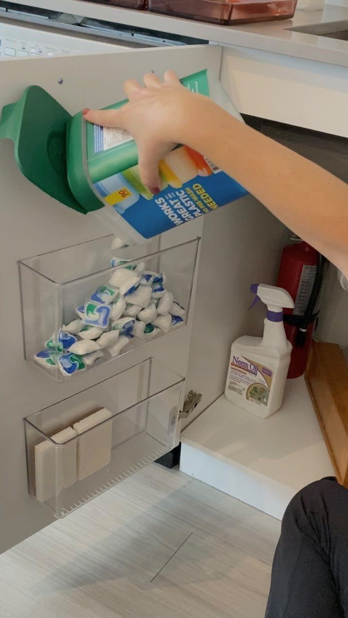 A hand reaches for plastic wrap stored in a clear organizer mounted on a cabinet door.