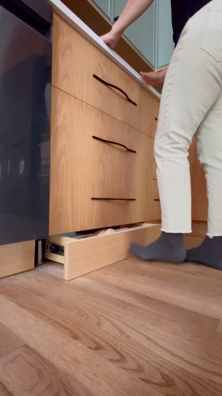 A foot pushes open a wooden toe kick drawer at the base of a kitchen cabinet.