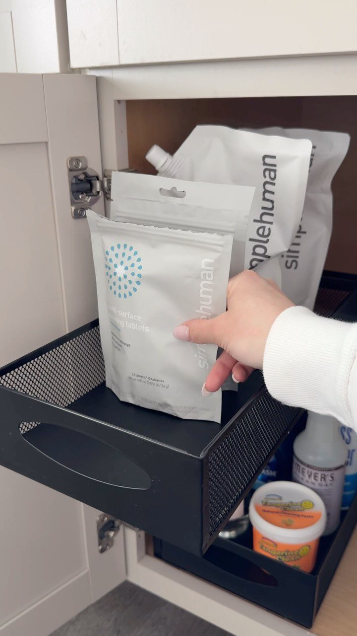 A hand reaches for a white pouch of cleaning wipes inside a black wire cabinet organizer.