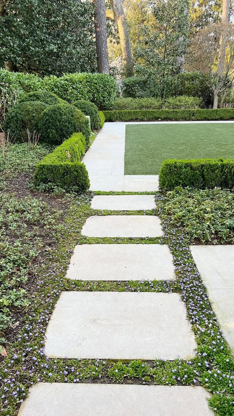 Rectangular stone pavers set in a lush garden with small purple flowers growing in the crevices.