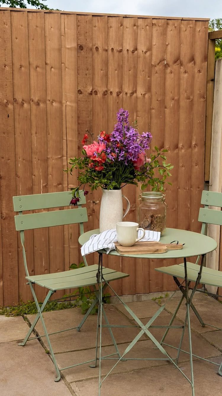 Sage green metal bistro set with flowers and a mug on a wooden patio.