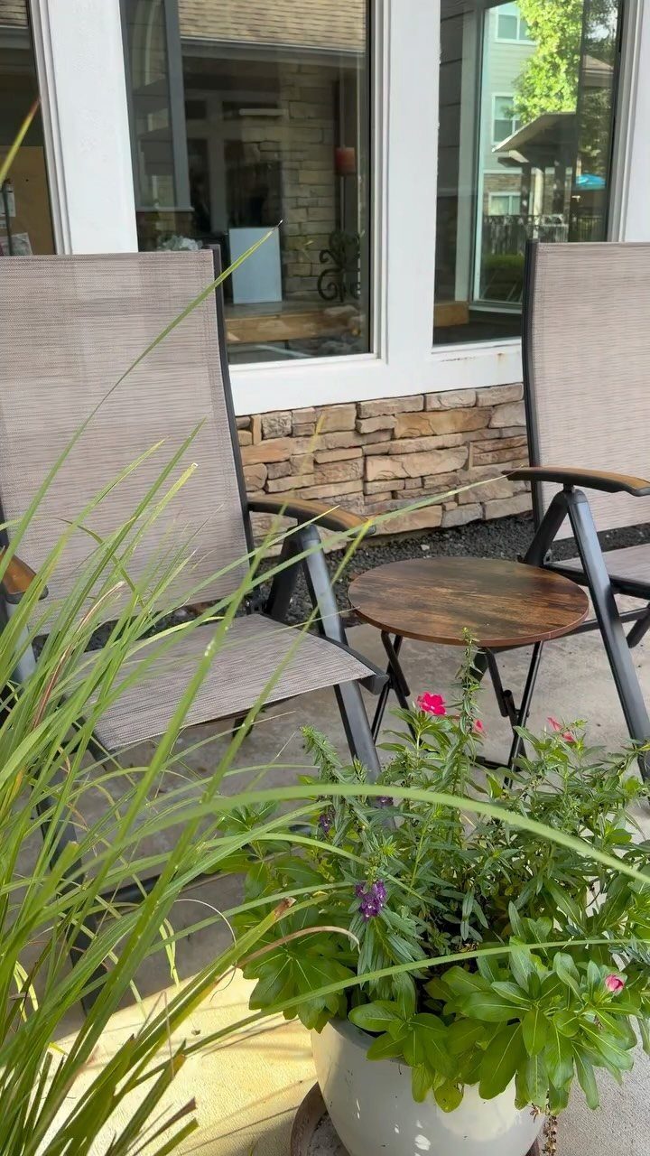 Two grey sling chairs and a small wooden table positioned near green plants on a patio.