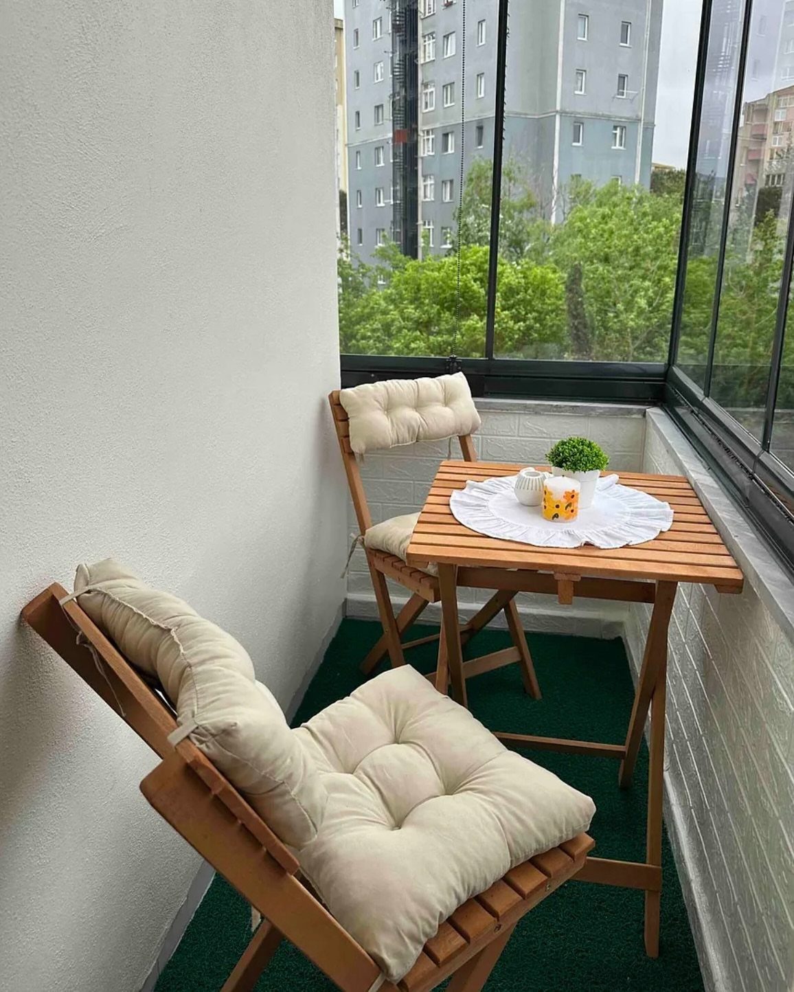 Two wooden chairs with cream cushions and a small wooden table on a green balcony.