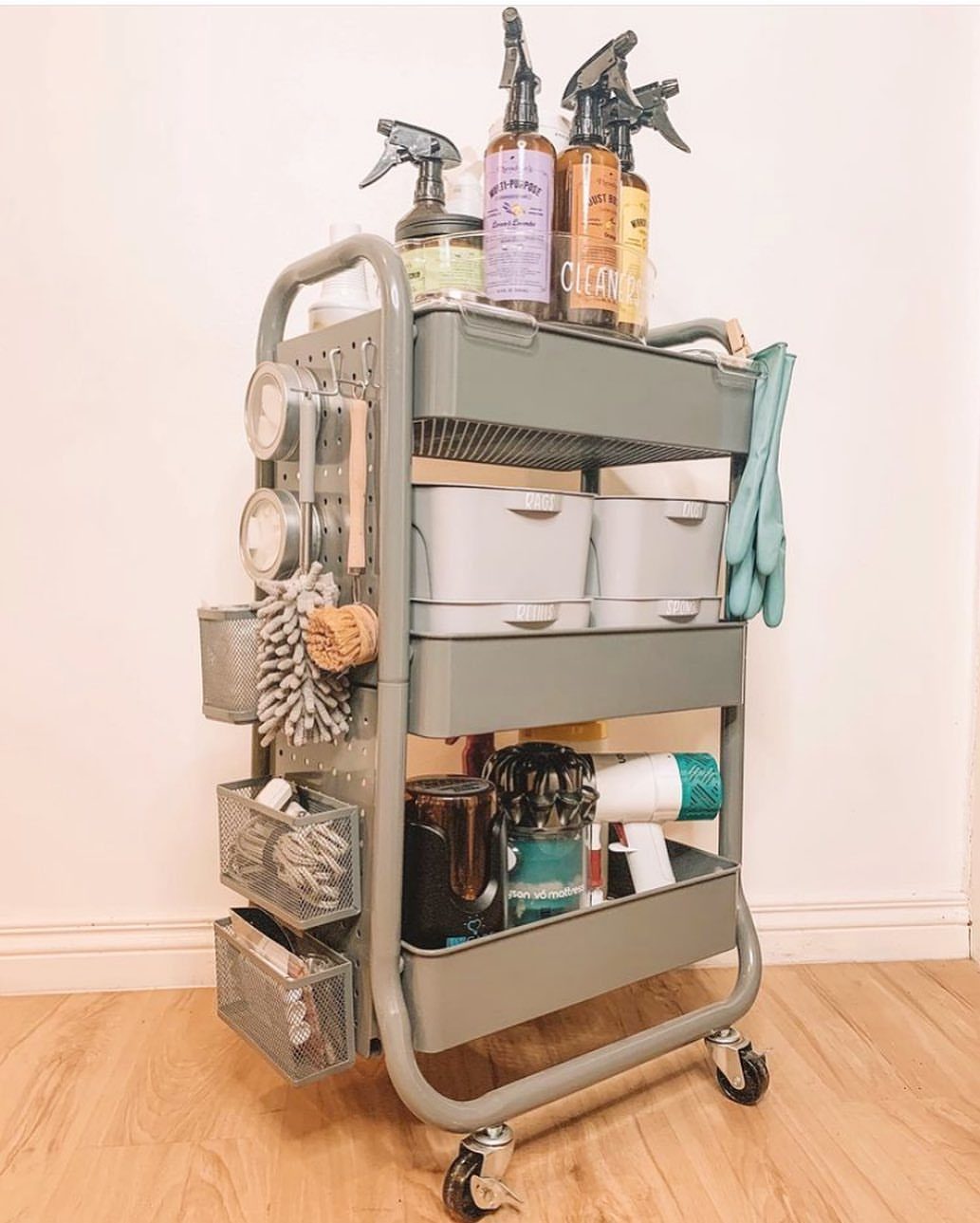 Grey three-tier rolling utility cart stocked with various cleaning supplies and spray bottles.