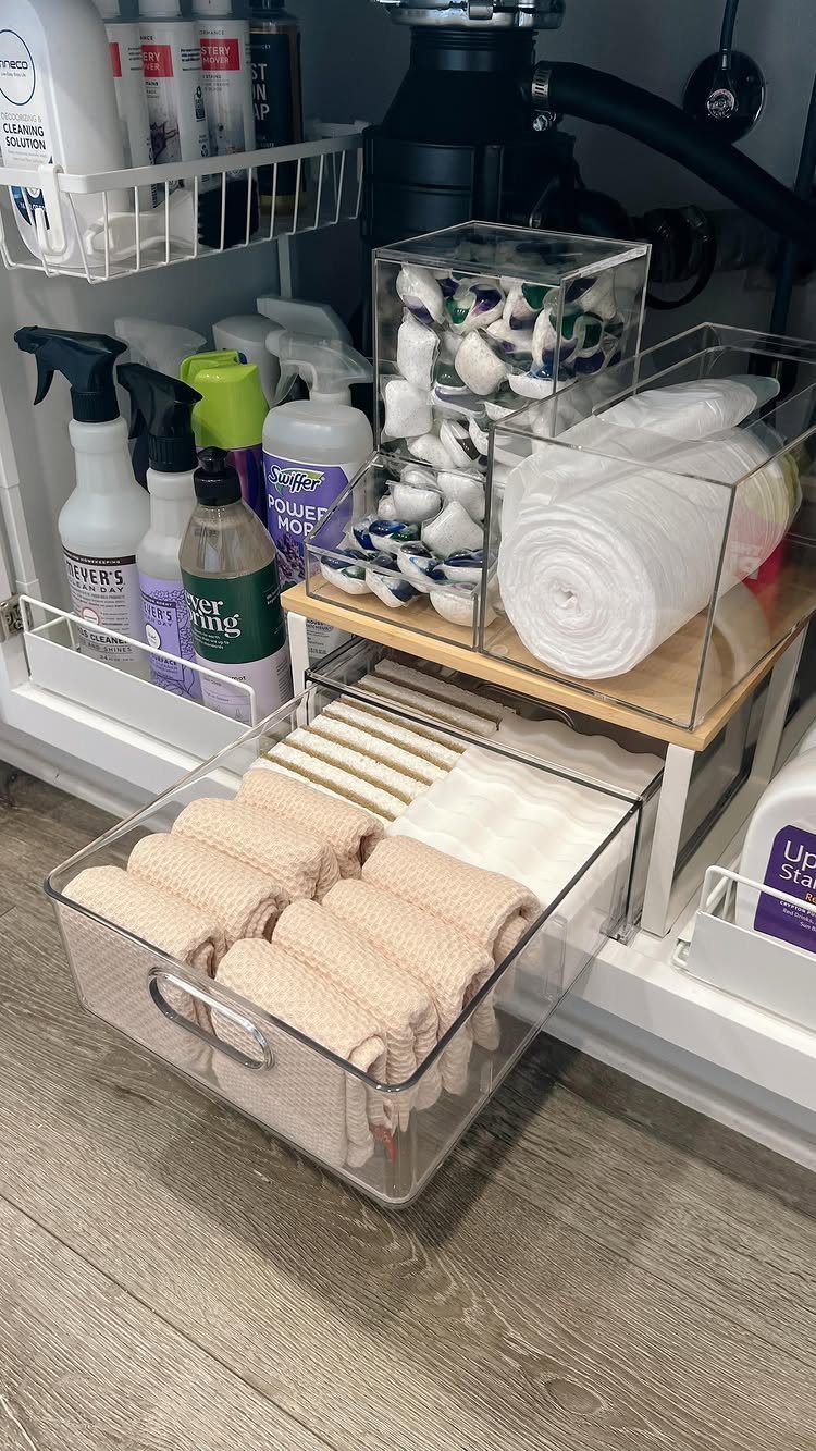 Organized cleaning supplies and rolled towels in clear plastic bins under a white sink.