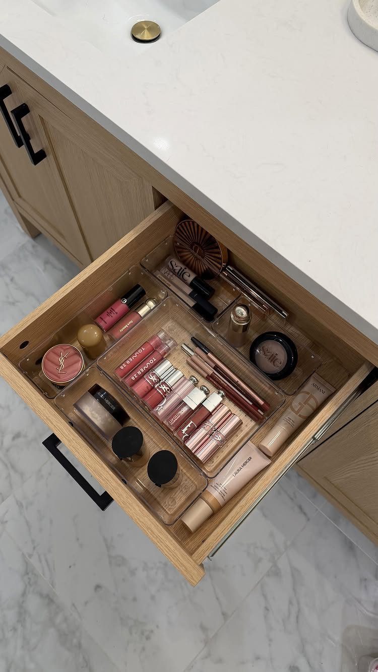 Clear acrylic organizers filled with various makeup products inside an open wooden vanity drawer.