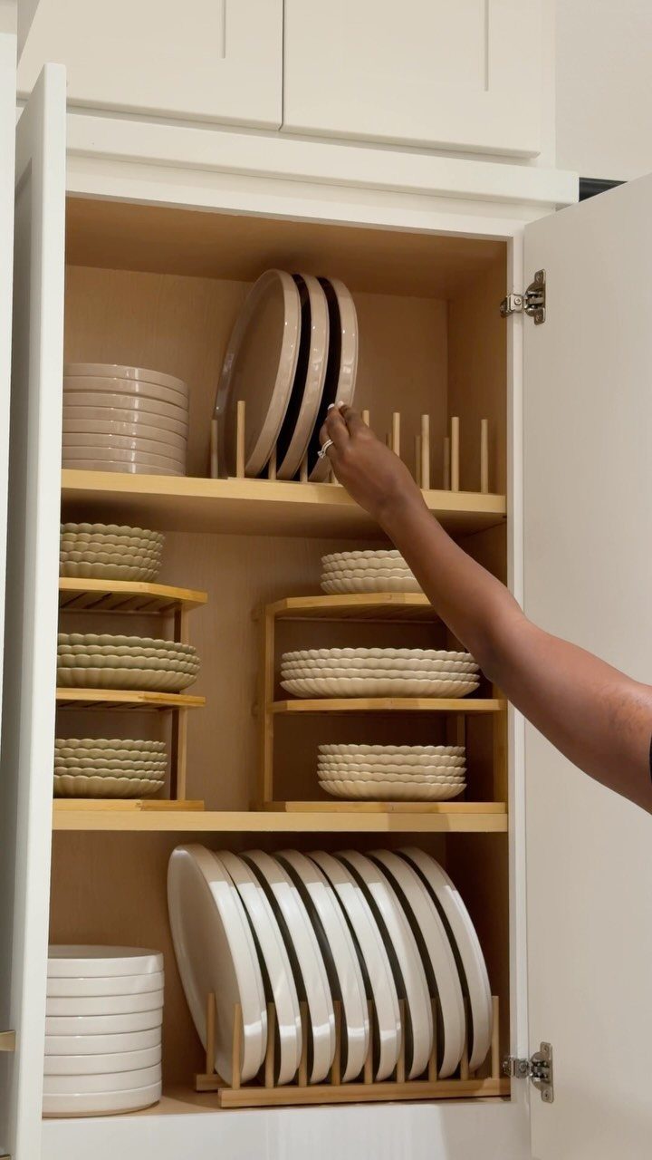 A hand reaching into a white kitchen cabinet to organize white plates on wooden shelf organizers.