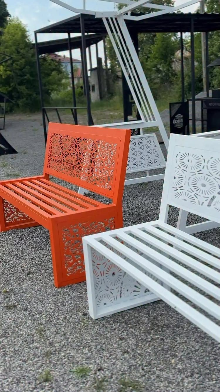 An orange and a white laser-cut metal bench sit on a gravel ground outdoors.