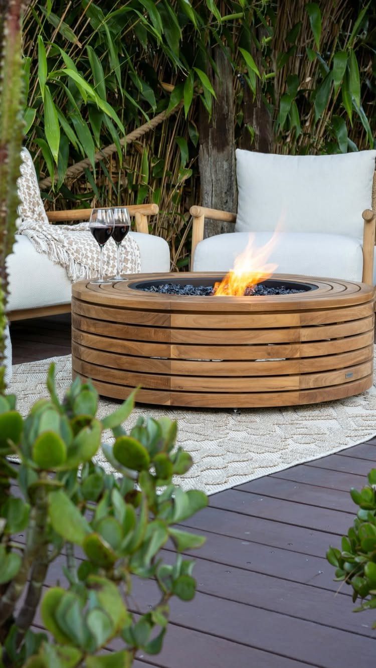 A circular wooden fire table with a bright flame sits on a deck between white lounge chairs.