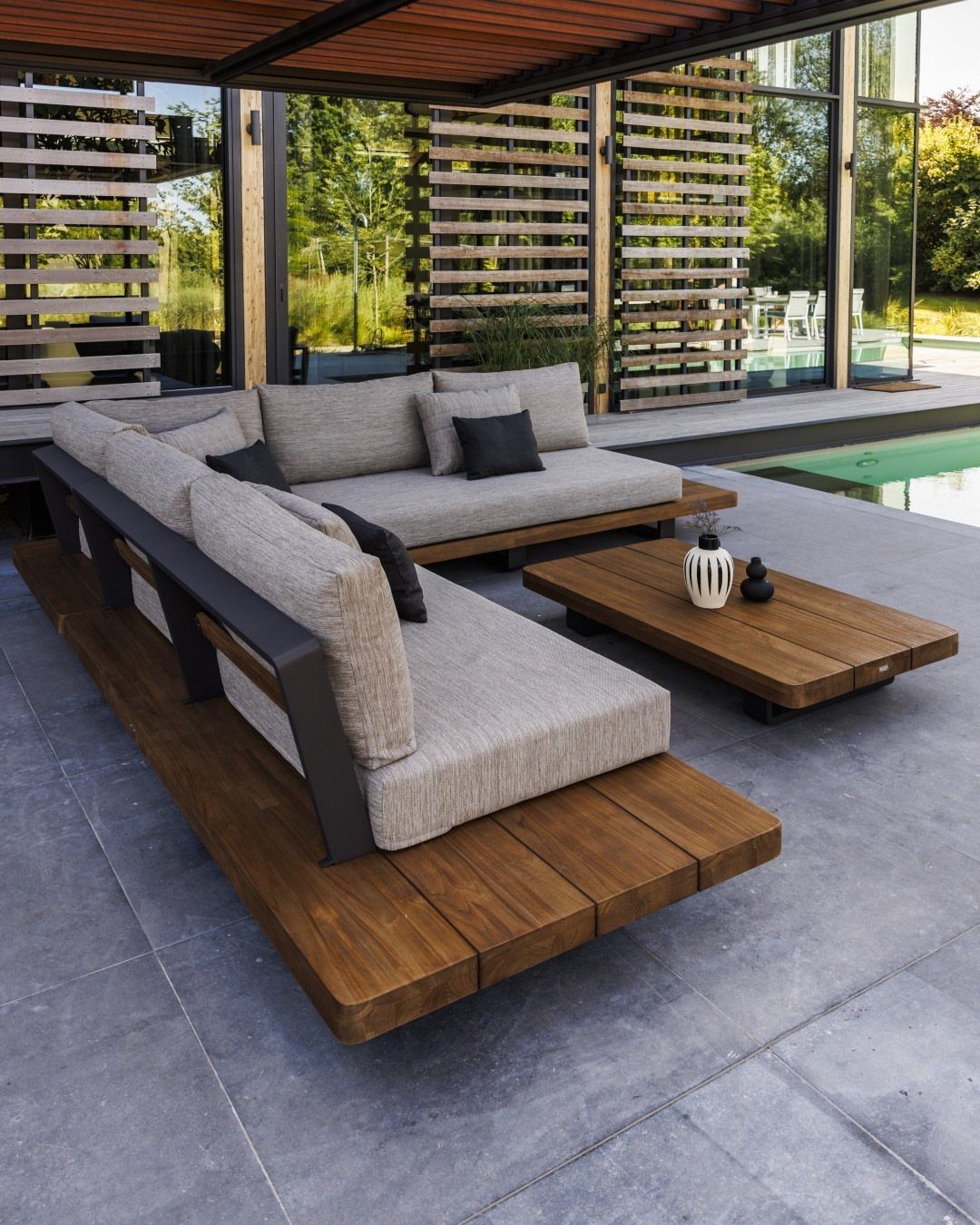 Modern outdoor lounge furniture with grey cushions and wooden platforms on a stone patio.