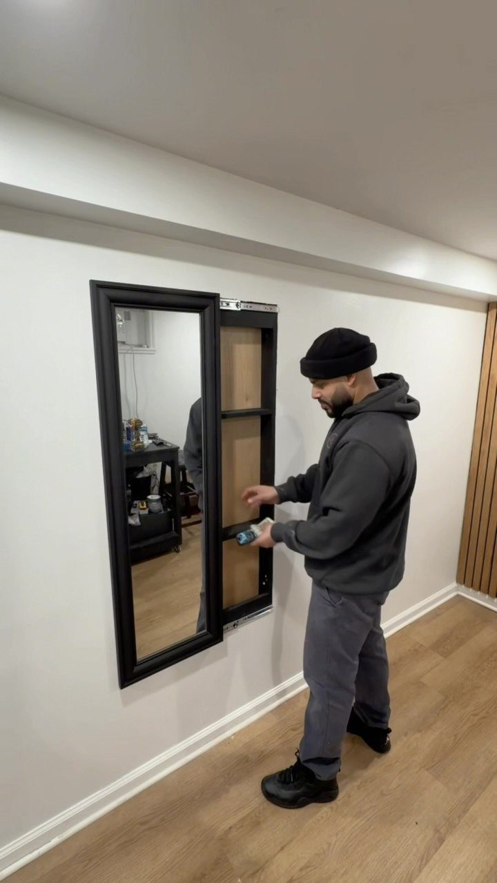 A man opens a large black-framed mirror to reveal a hidden wooden shelf compartment.