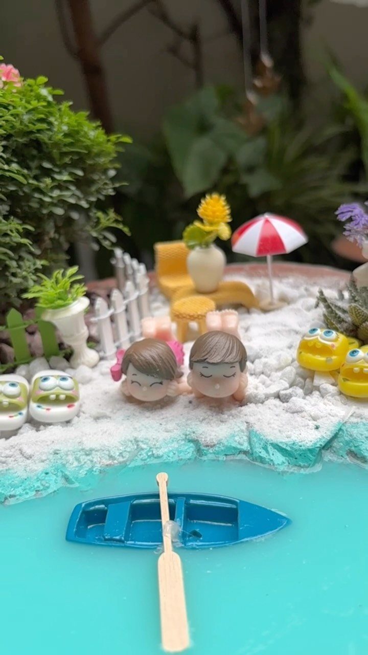 23 Fairy Garden Ideas for Tiny Houses, Teacups and Tree Trunks 3 Miniature beach scene with two small figures, a tiny blue boat, and a striped umbrella on white sand.