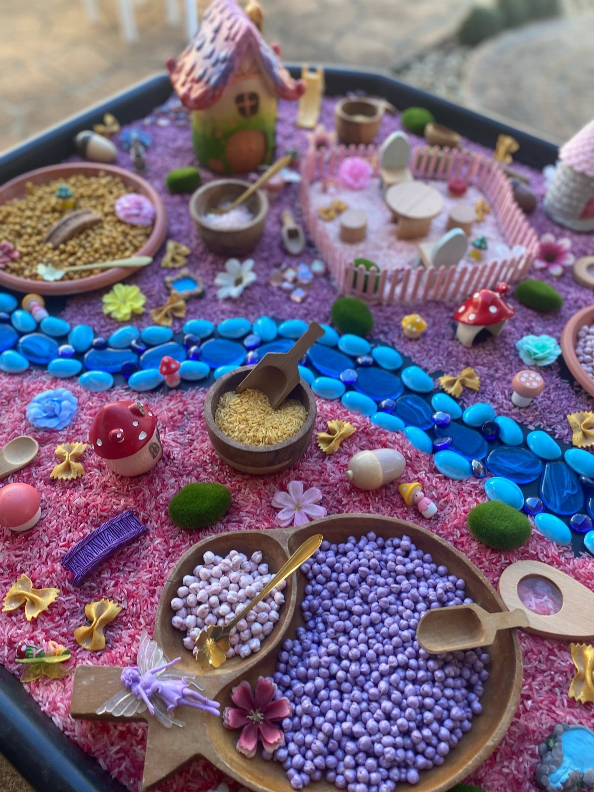 23 Fairy Garden Ideas for Tiny Houses, Teacups and Tree Trunks 18 Sensory bin with pink base, blue pebbles, miniature mushrooms, and small wooden bowls of beads.