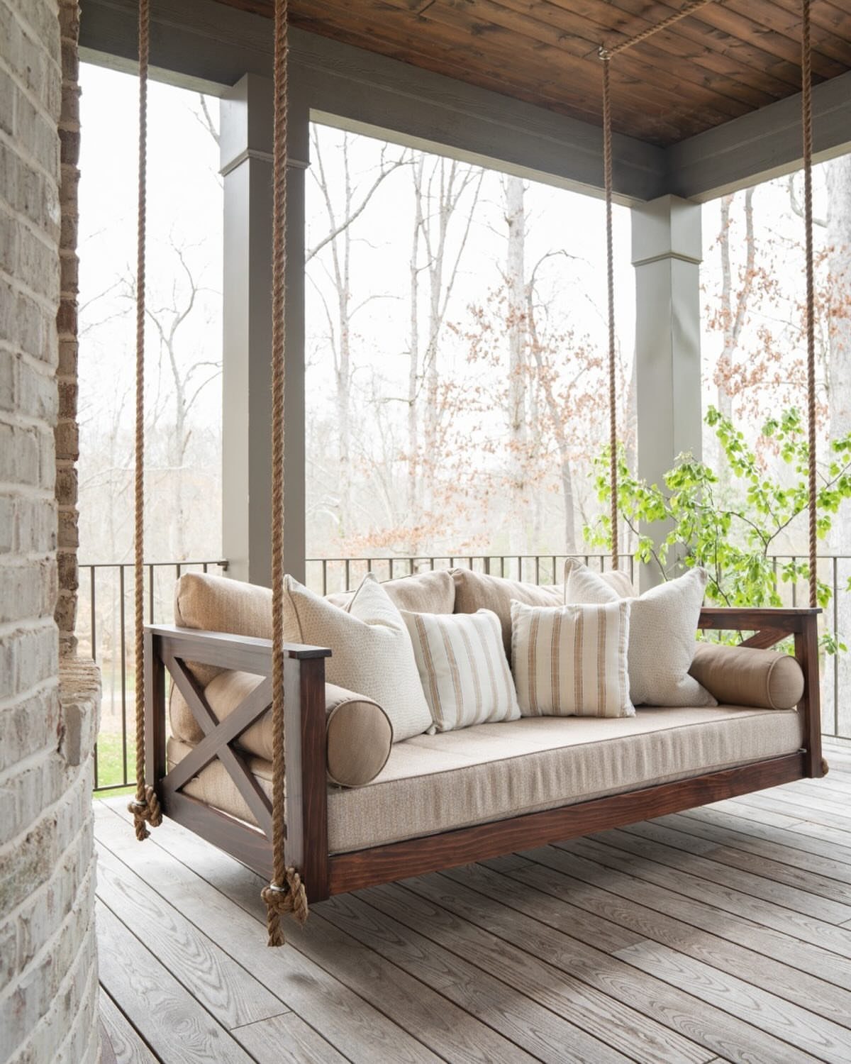 14 Front Porch Decorating Ideas for Every Season 10 A wooden porch swing with neutral cushions and pillows hanging by ropes on a porch.