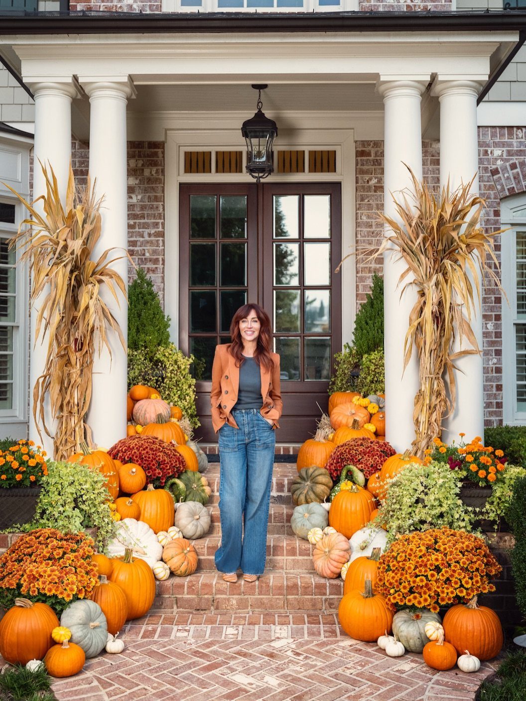 14 Front Porch Decorating Ideas for Every Season 11 Woman standing on a brick porch decorated with pumpkins, gourds, and tall corn stalks.