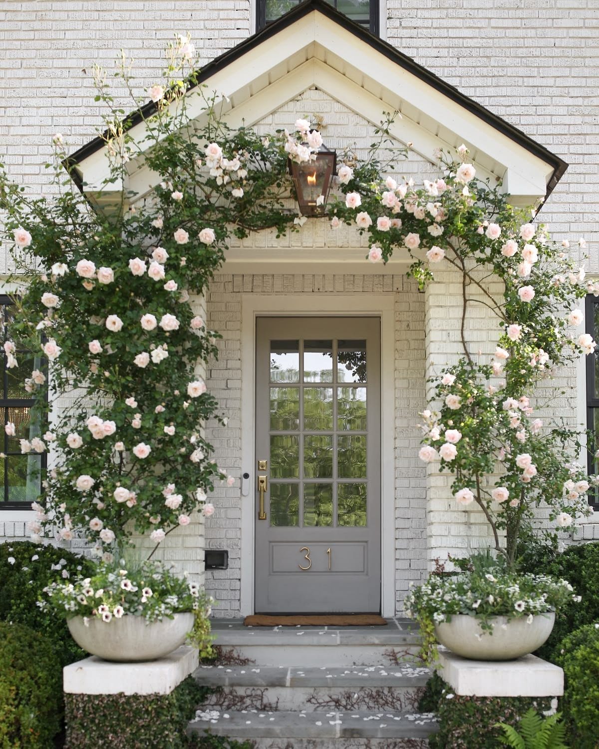 14 Front Porch Decorating Ideas for Every Season 12 Climbing pink roses arching over a white brick house entrance with grey door and stone planters.