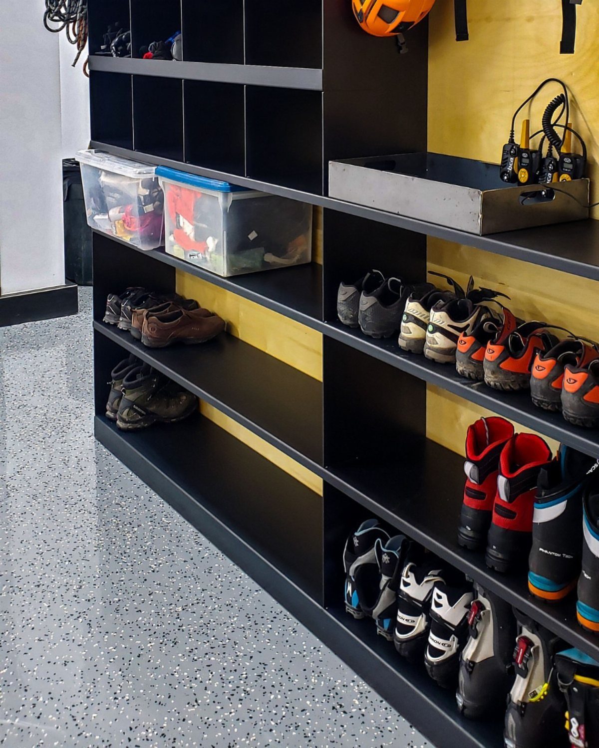 9 Garage Storage Shoes For Any Budget, Wall to Floor 8 Black garage shelving units holding storage bins and various pairs of shoes against a yellow wall.