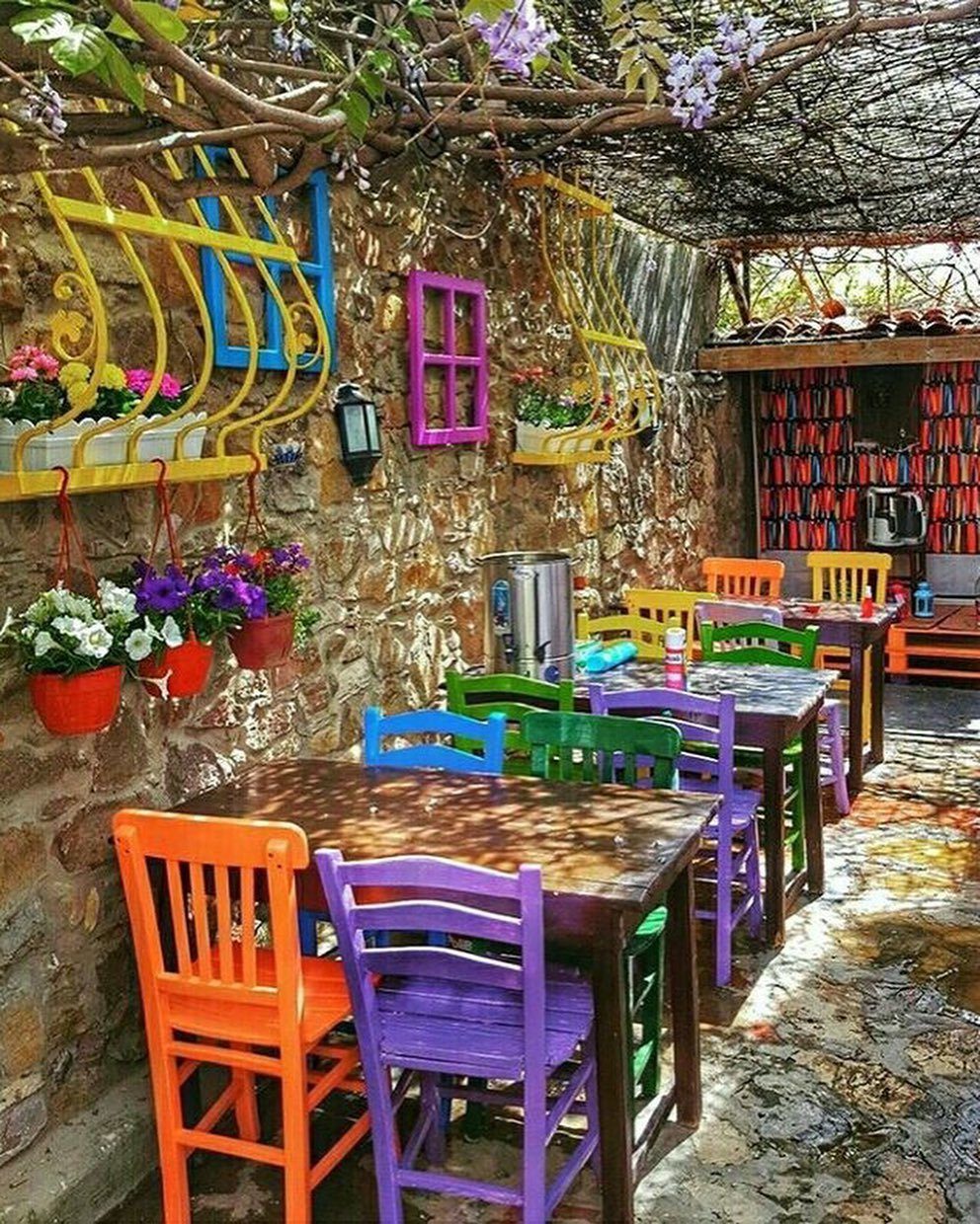 Brightly colored wooden chairs in orange, purple, and green arranged around tables in a stone patio garden.