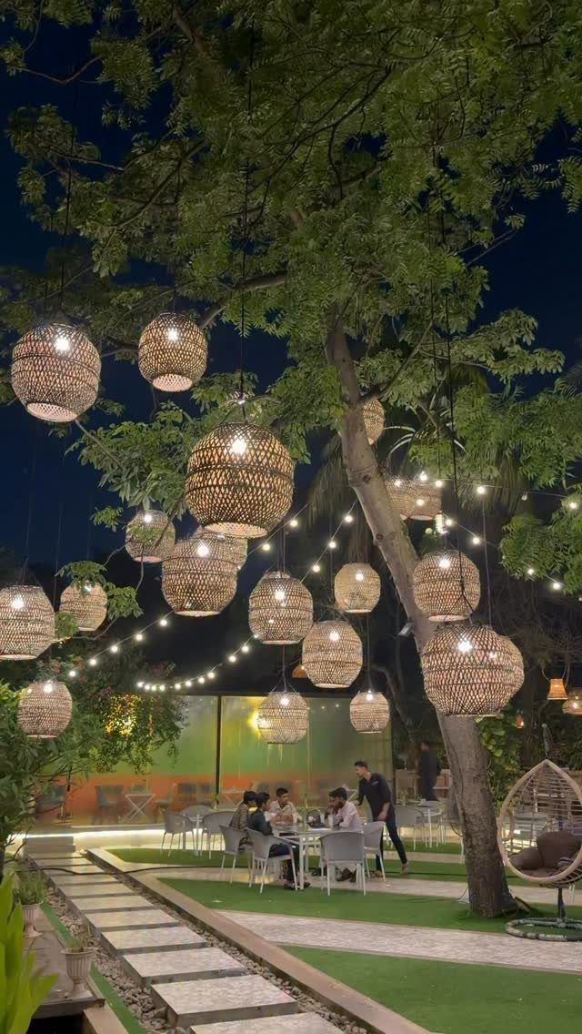 Woven wicker spherical lanterns hanging from tree branches in a lit outdoor garden setting at night.