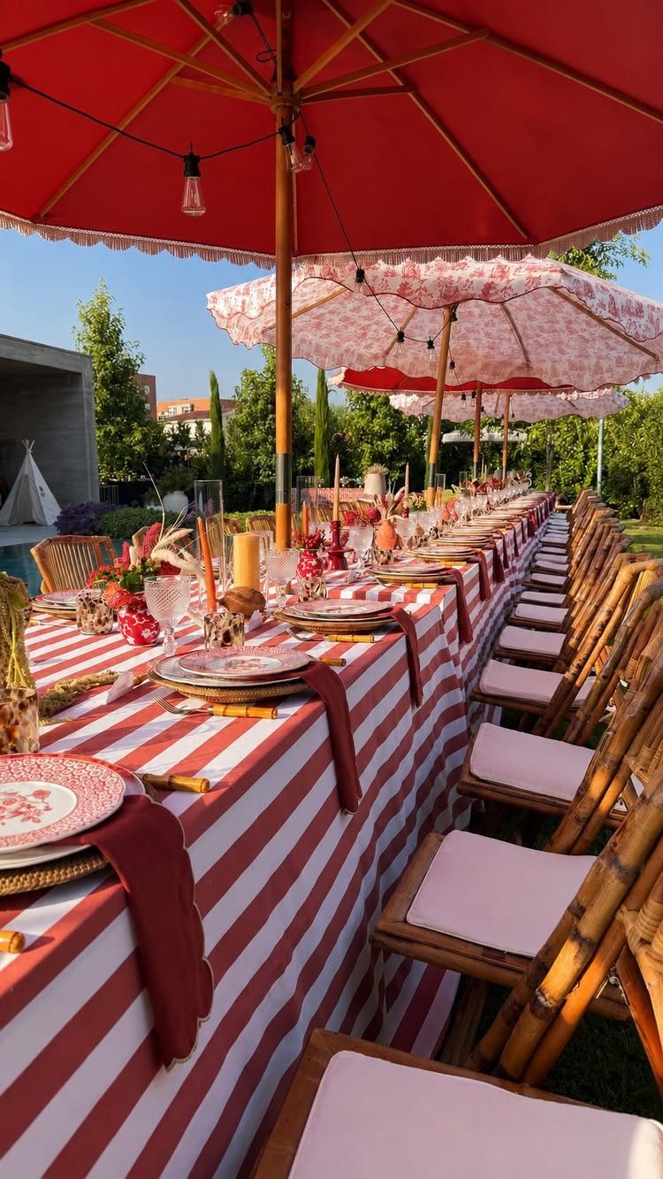 A long dining table decorated with red and white striped linens and large red umbrellas outdoors.