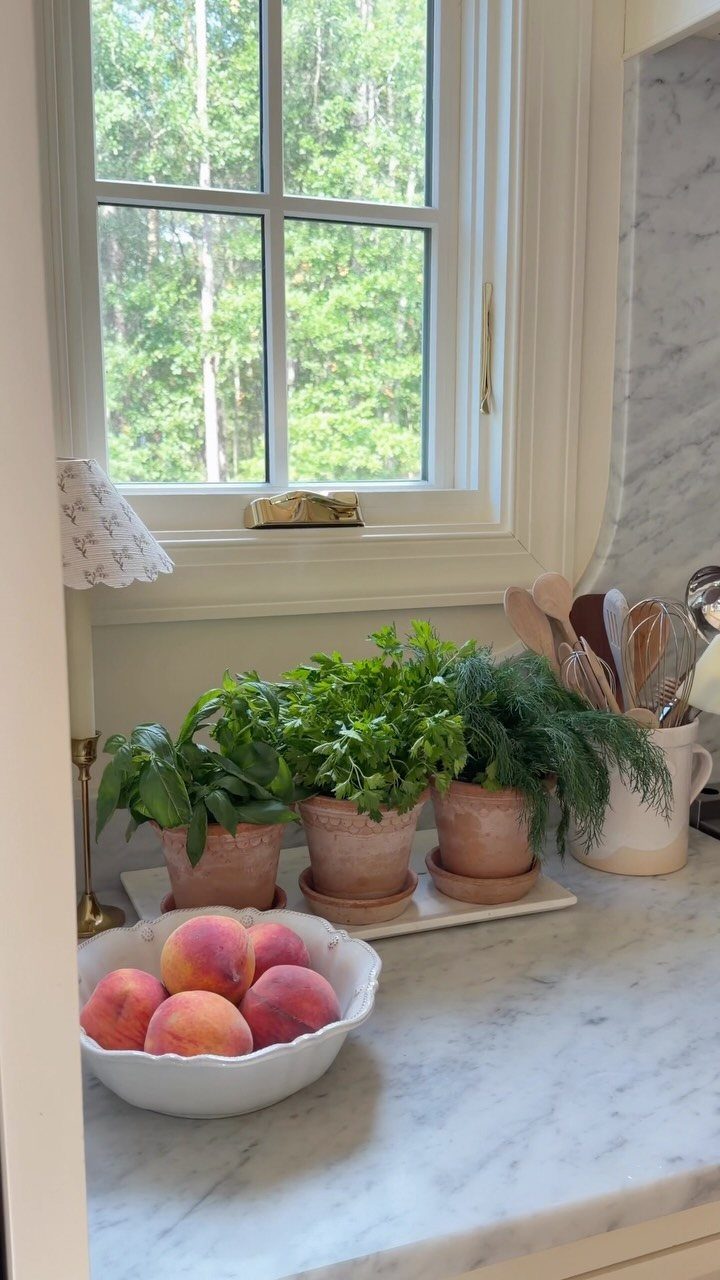 Three terracotta pots with green herbs sit on a marble kitchen countertop near a window.