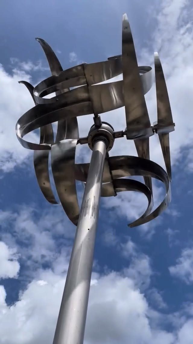Metal Yard Art Ideas for Your Garden in 7 Styles 2 Stainless steel abstract sculpture with curved metal blades mounted on a tall pole against a cloudy sky.