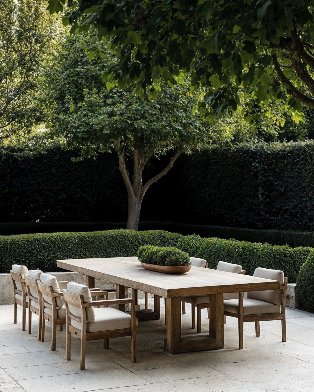 Patio Dining Table and 7 Outdoor Eating Area Ideas 2026 2 Large wooden dining table with eight cushioned chairs set on a stone patio surrounded by greenery.