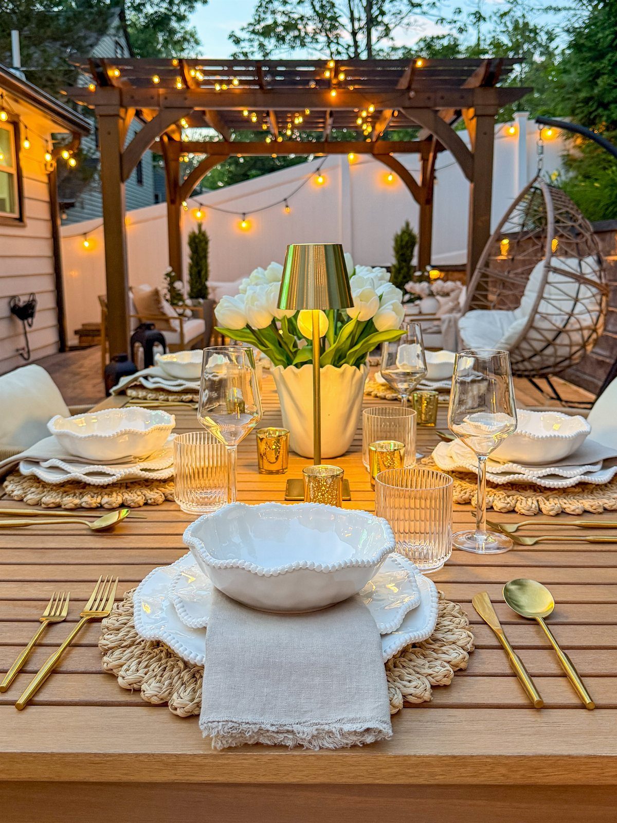 Patio Dining Table and 7 Outdoor Eating Area Ideas 2026 3 A wooden dining table set with white bowls, gold utensils, and white tulips under a lit pergola.