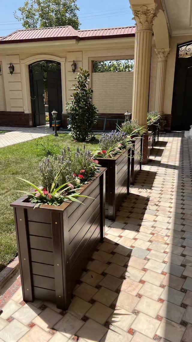 13 Planter Boxes Ideas for Your Porch, Deck and Garden 13 Dark brown wooden planter boxes with green plants and purple flowers on a tiled walkway.