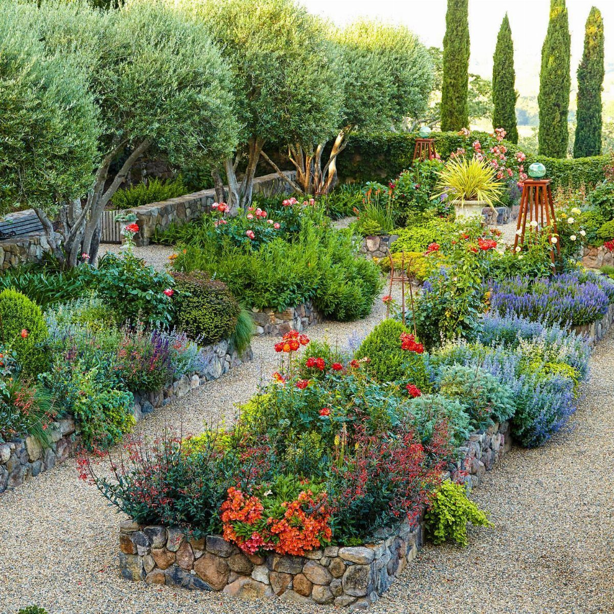 14 Raised Garden Bed Ideas for Cedar, Stone, Metal & More 11 Stone raised garden beds filled with colorful flowers in a gravel courtyard with olive trees.