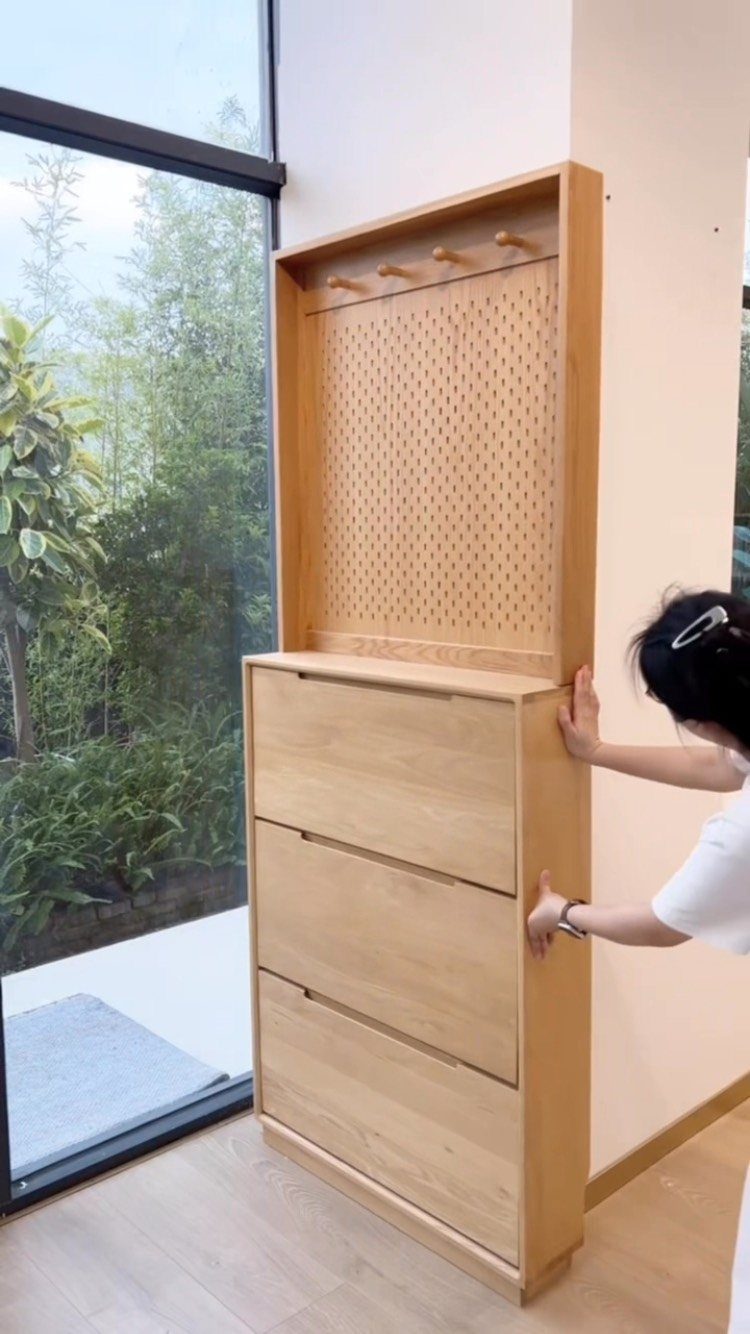 A person opening a large drawer of a tall wooden shoe cabinet near a window.