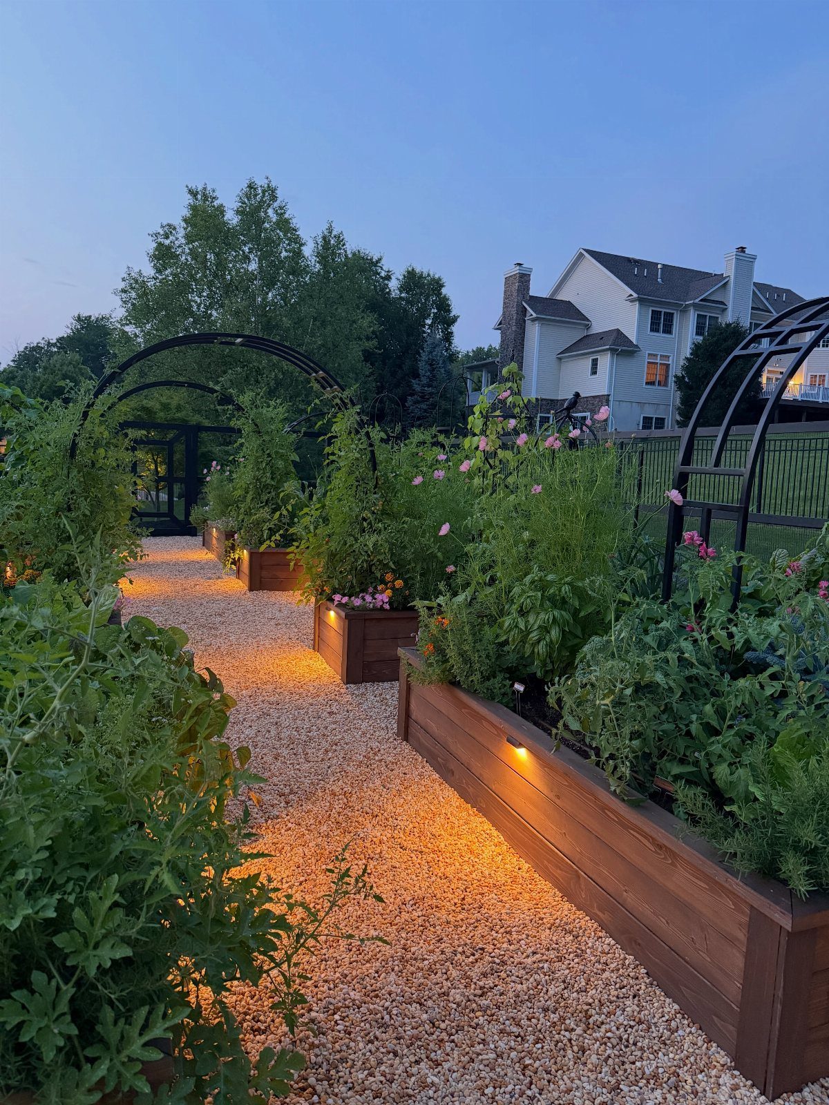 18 Small Backyard Garden Ideas for Compact Outdoor Style 3 A lit gravel pathway winding between wooden raised garden beds under black metal arches.