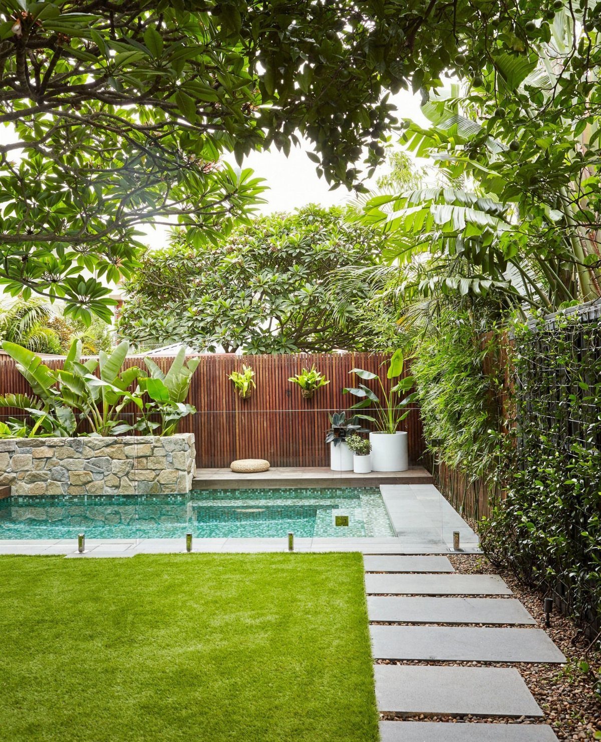 18 Small Backyard Garden Ideas for Compact Outdoor Style 9 A backyard with a swimming pool, green lawn, stone pavers, and a wooden privacy fence.