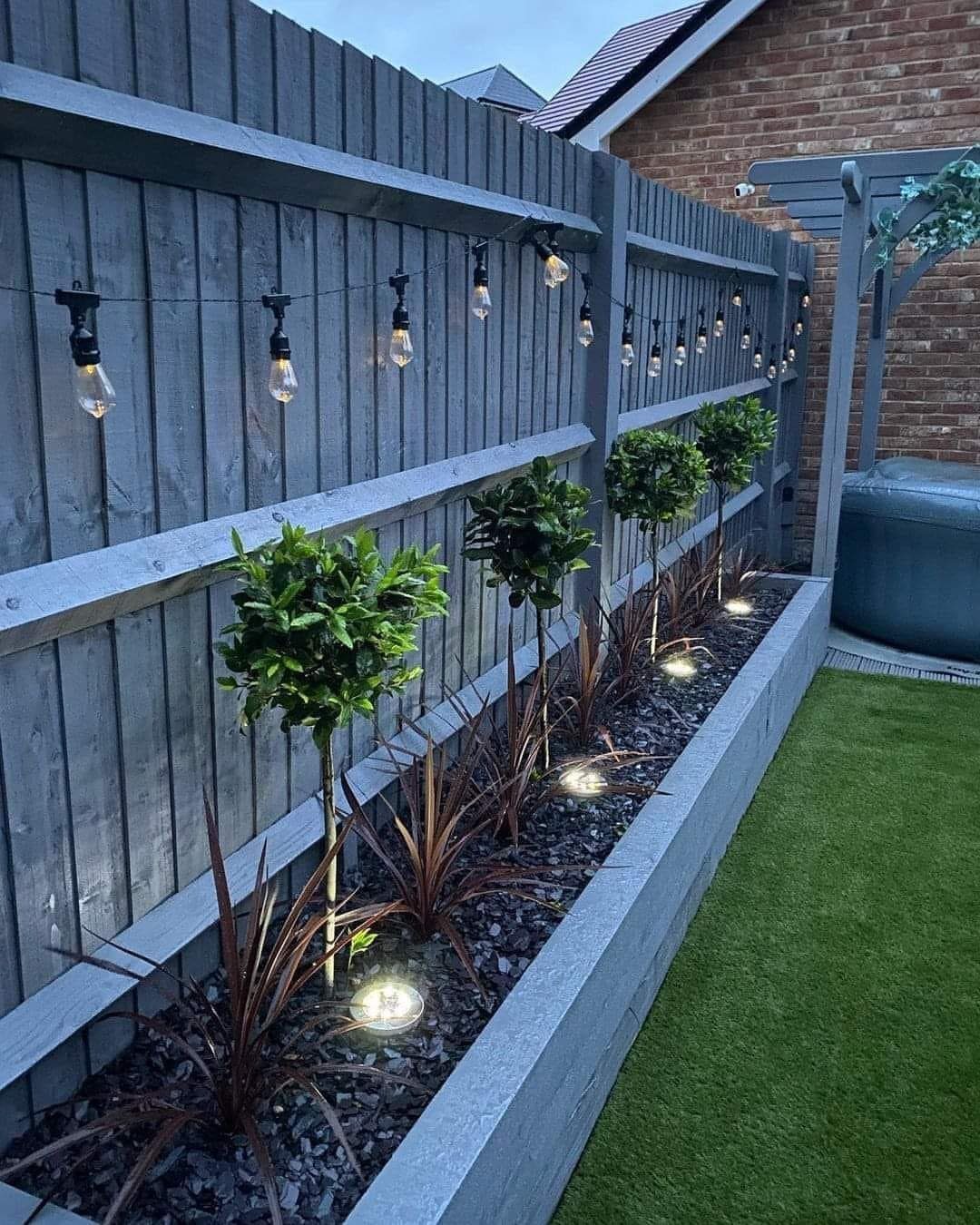 18 Small Backyard Garden Ideas for Compact Outdoor Style 4 String lights and ground spotlights illuminate small trees and plants in a narrow garden bed.