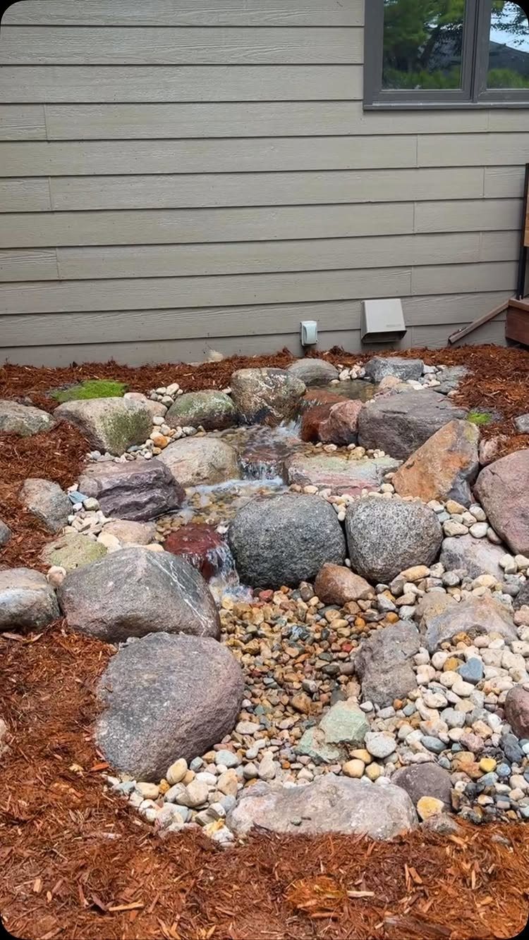 Small rock stream water feature flowing through pebbles and mulch in a backyard garden.