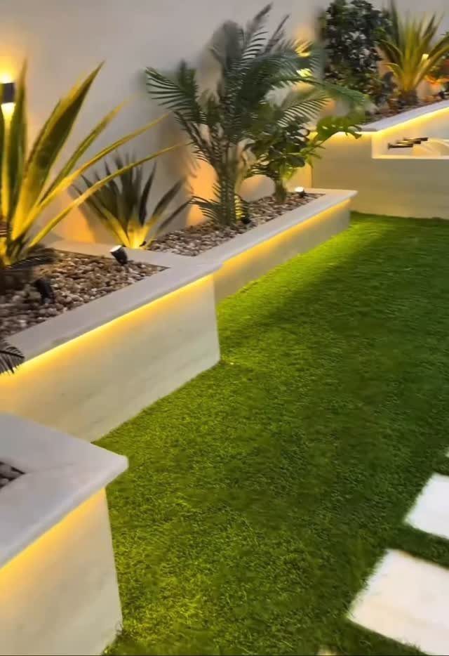 Modern landscaped yard with artificial grass, tropical plants in white stone planters, and warm LED lighting.