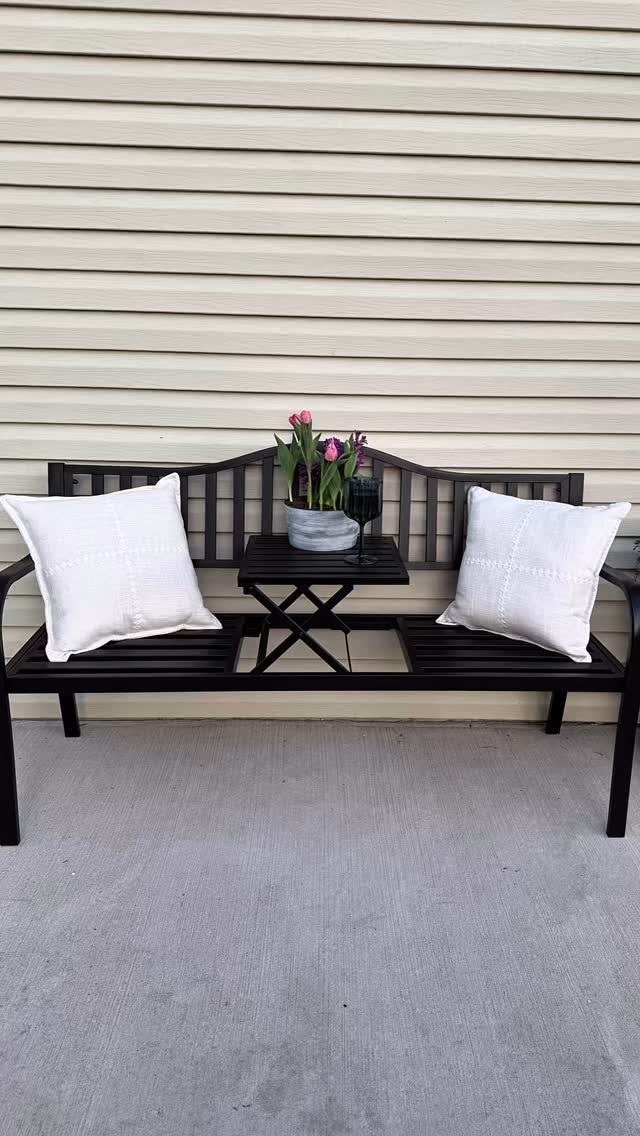 Black metal garden bench with white pillows and a small side table against beige siding.