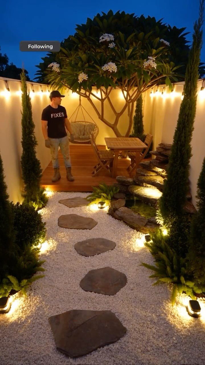 A small landscaped backyard with a wooden deck, stone path, and illuminated greenery at twilight.