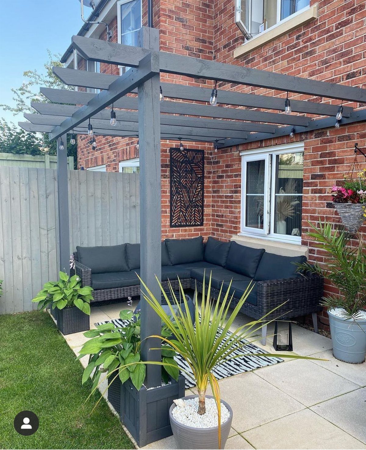Grey wooden pergola over a patio with wicker seating and potted plants against a brick wall.