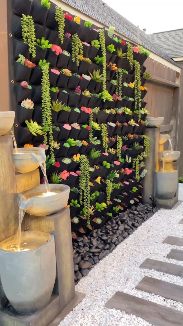 A black vertical wall planter filled with colorful succulents next to tiered stone water fountains.