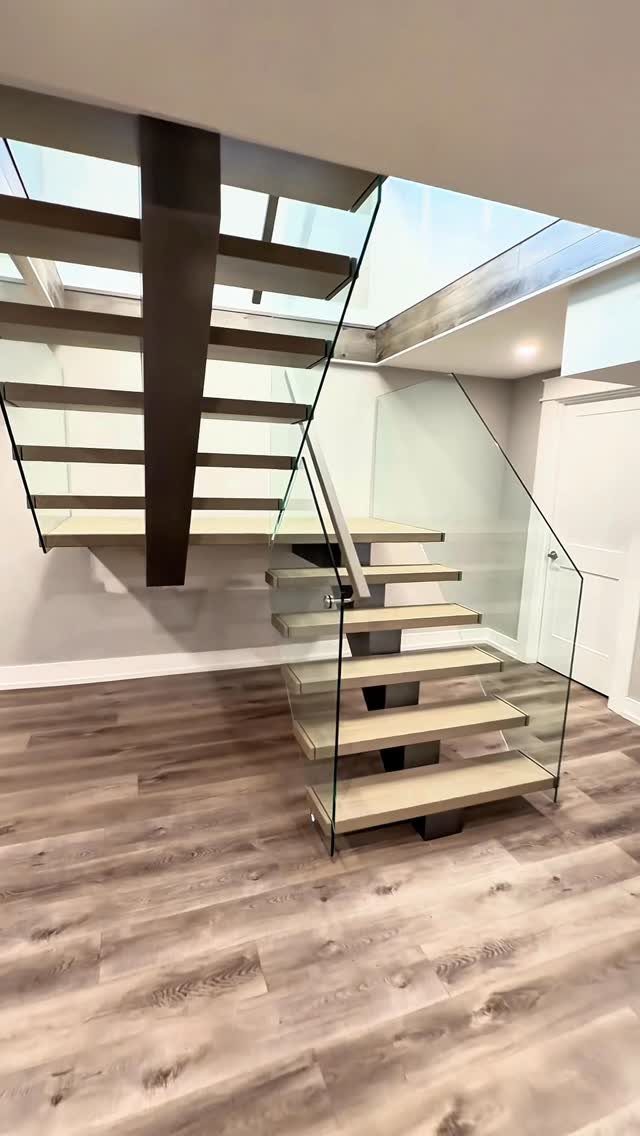 Floating stairs with light wood treads, a dark metal support, and clear glass railings on wood flooring.