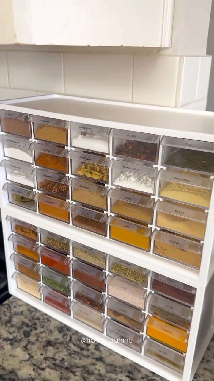 A white tiered organizer filled with various colorful spices in clear plastic drawers.