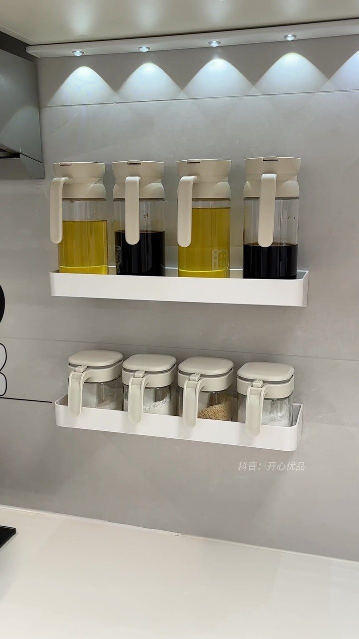 Two white wall-mounted shelves holding glass jars of oils and spices against a grey wall.